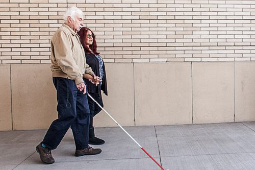 A woman holding the hand of an elderly blind man while walking down the sidewalk.